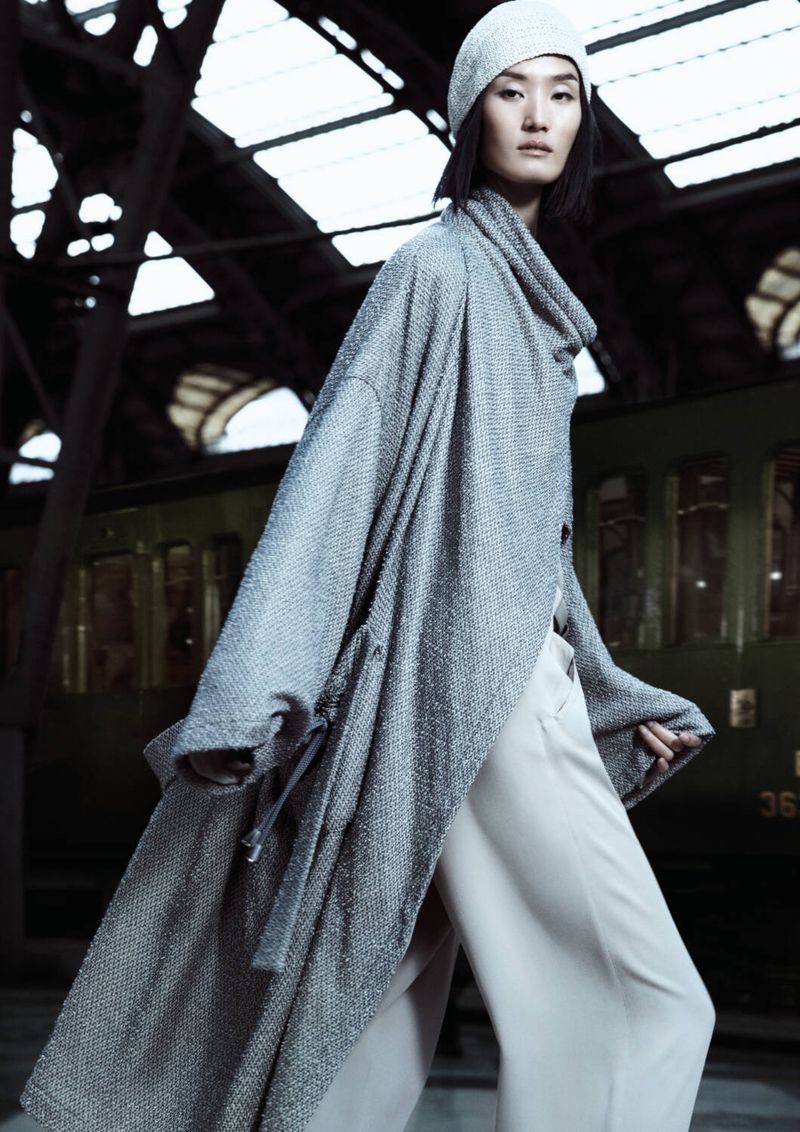 A woman poses in a long gray coat and hat on a train platform. The photo is in portrait orientation and showcases fashion apparel.