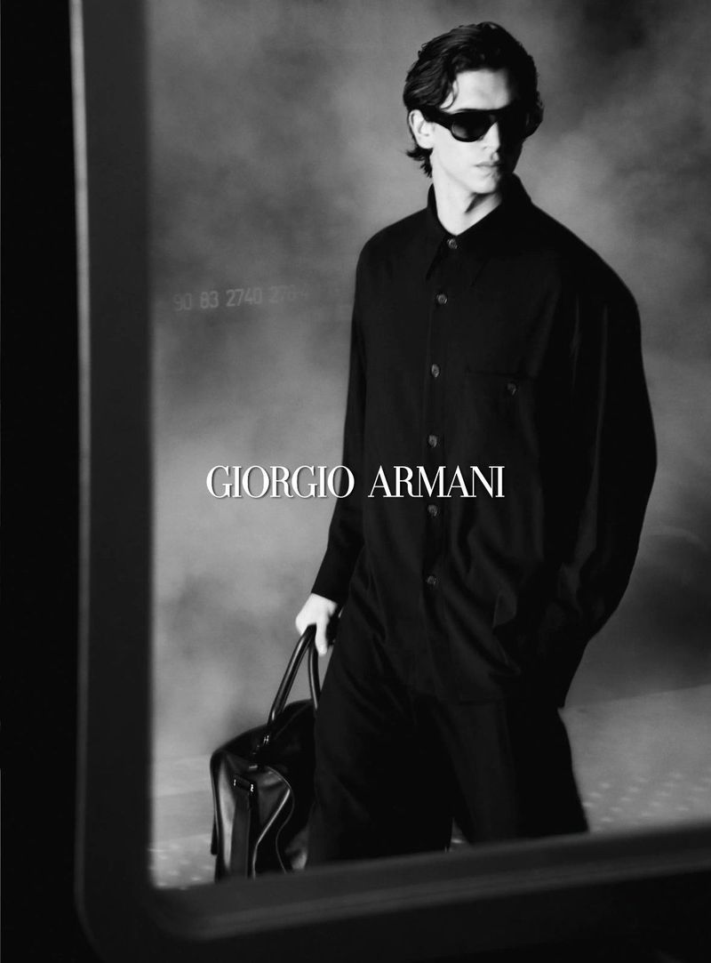 Black and white fashion shot of a man in sunglasses and all-black outfit carrying a duffel bag, showcasing Giorgio Armani brand.