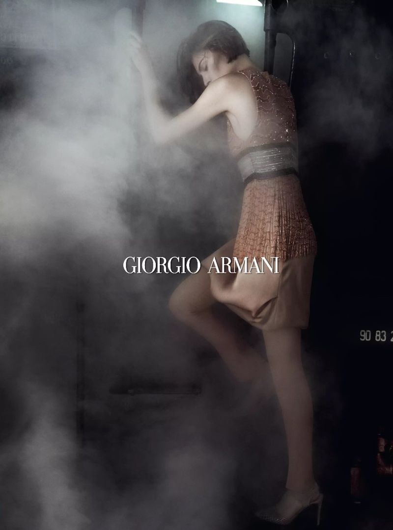 A woman in a short embellished dress poses in a dark, foggy environment, featuring the Giorgio Armani logo and highlighting fashion and style.
