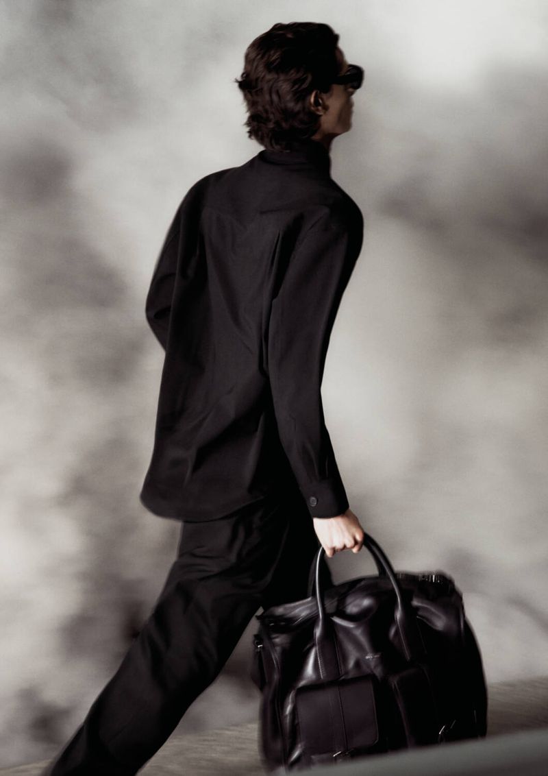 Adult male in a black suit carries a black leather bag against a blurred background, creating a stylish and fashionable outdoor portrait.
