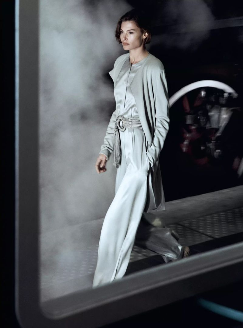 Fashionable adult woman strides confidently in light-blue pantsuit, cinched with an elaborate belt, against a backdrop of billowing smoke.