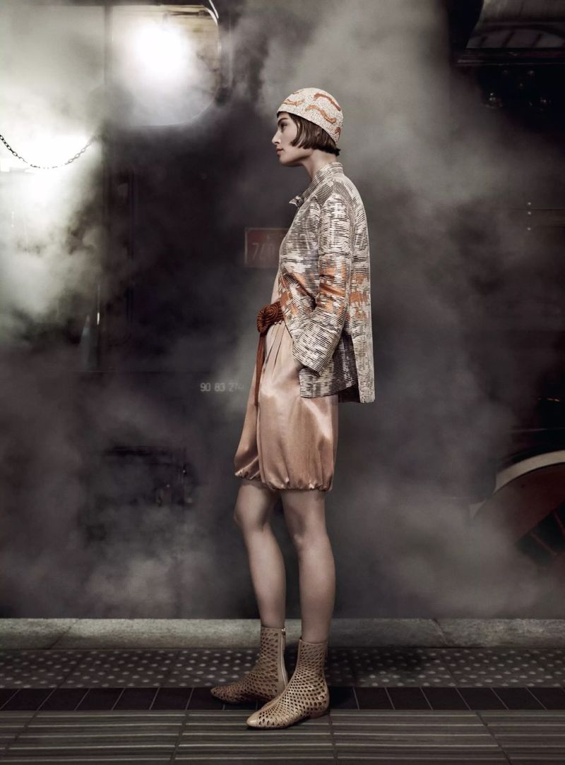 Woman in patterned jacket and hat stands on platform amid smoke. Side view of fashion shot in an industrial setting with tiled floor.