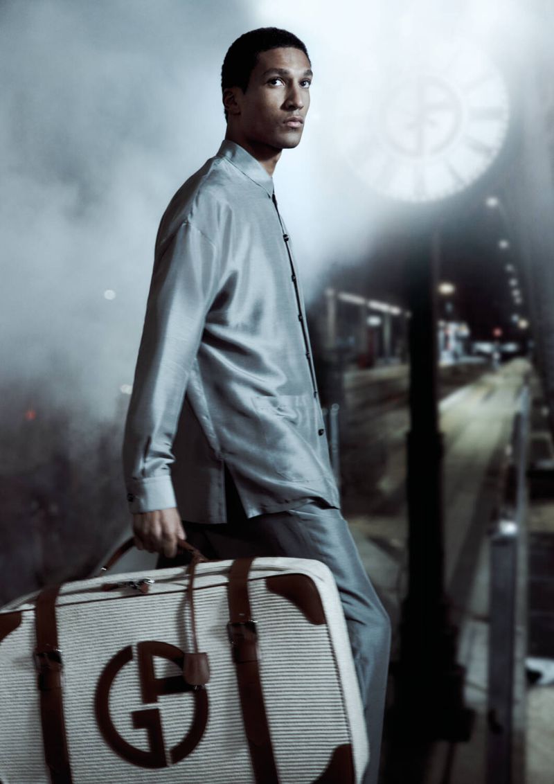 Man in suit at train station with Armani luggage. Male fashion model poses with designer bag at an outdoor railway platform.