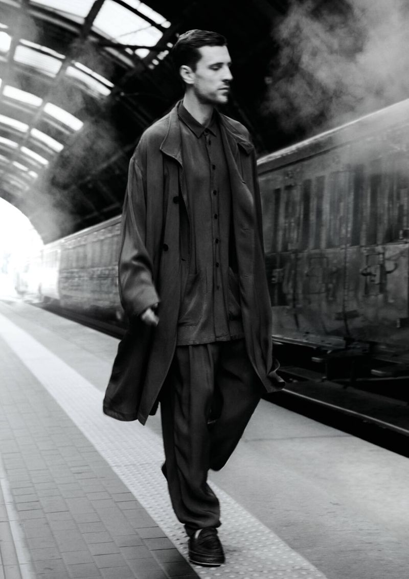 Man walking along a train platform wearing a long coat, matching shirt and pants, and sneakers, with a vintage train in the background.