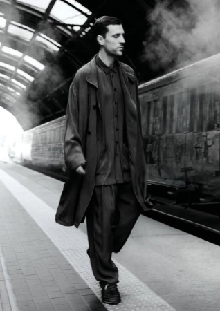 Man walking along a train platform wearing a long coat, matching shirt and pants, and sneakers, with a vintage train in the background.