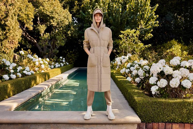 A woman in a tan quilted coat stands in front of a reflecting pool in a lush garden setting on a sunny day, looking directly at the camera.
