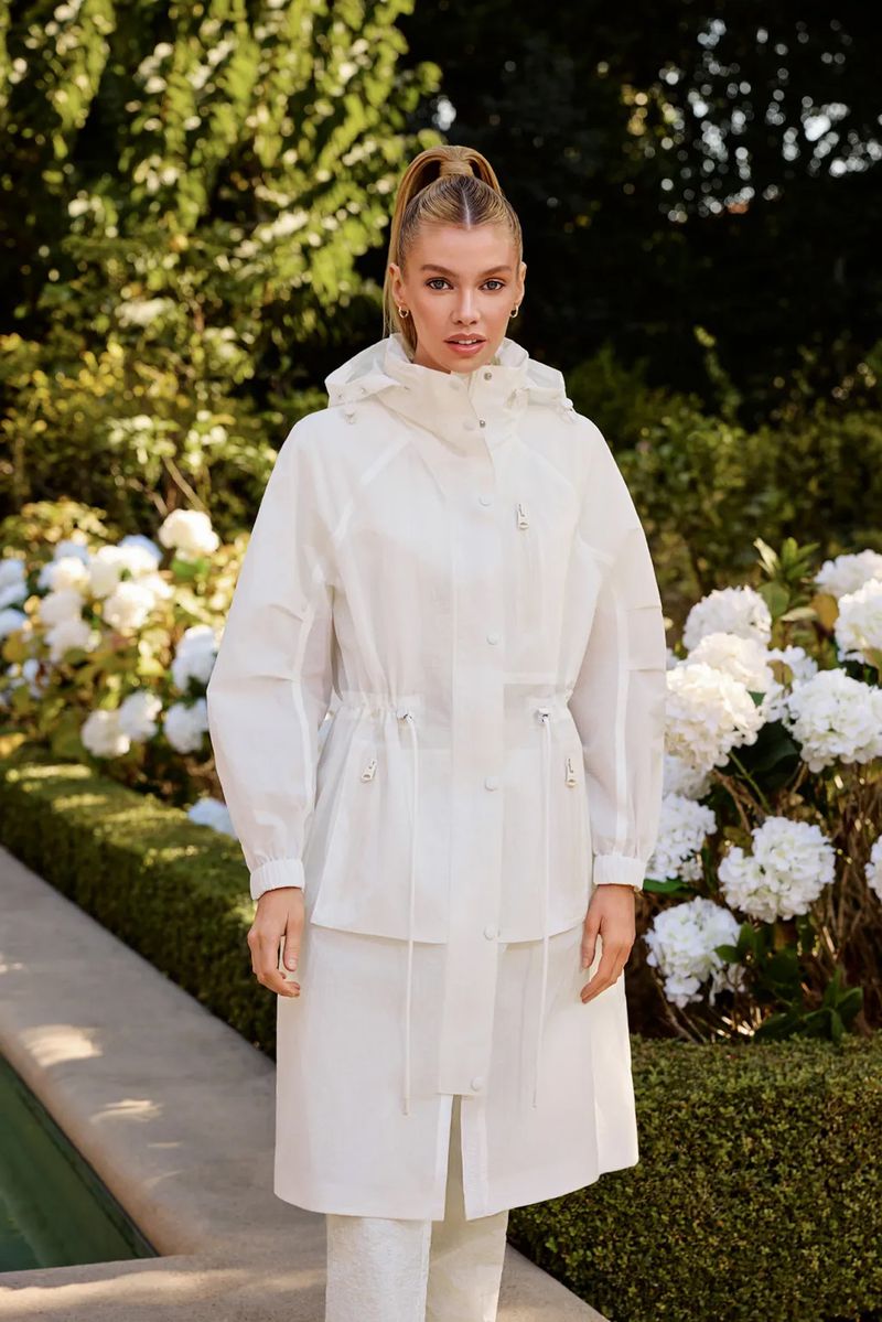 A stylish adult woman stands outdoors wearing a long white hooded raincoat with a cinched waist and light colored pants in a garden setting.