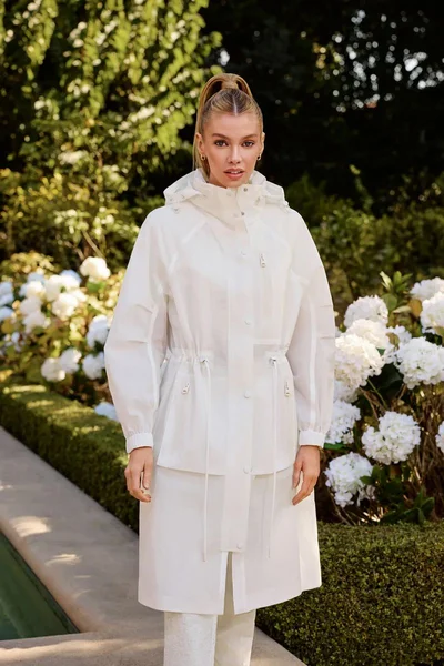Stella Maxwell in a white hooded utility coat outdoors by white hydrangea bushes for the Mackage S/S 2025 Campaign