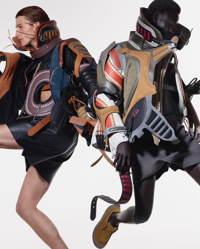Two adult fashion models strike dynamic poses in futuristic, deconstructed outfits against a bright white backdrop in a studio shot.