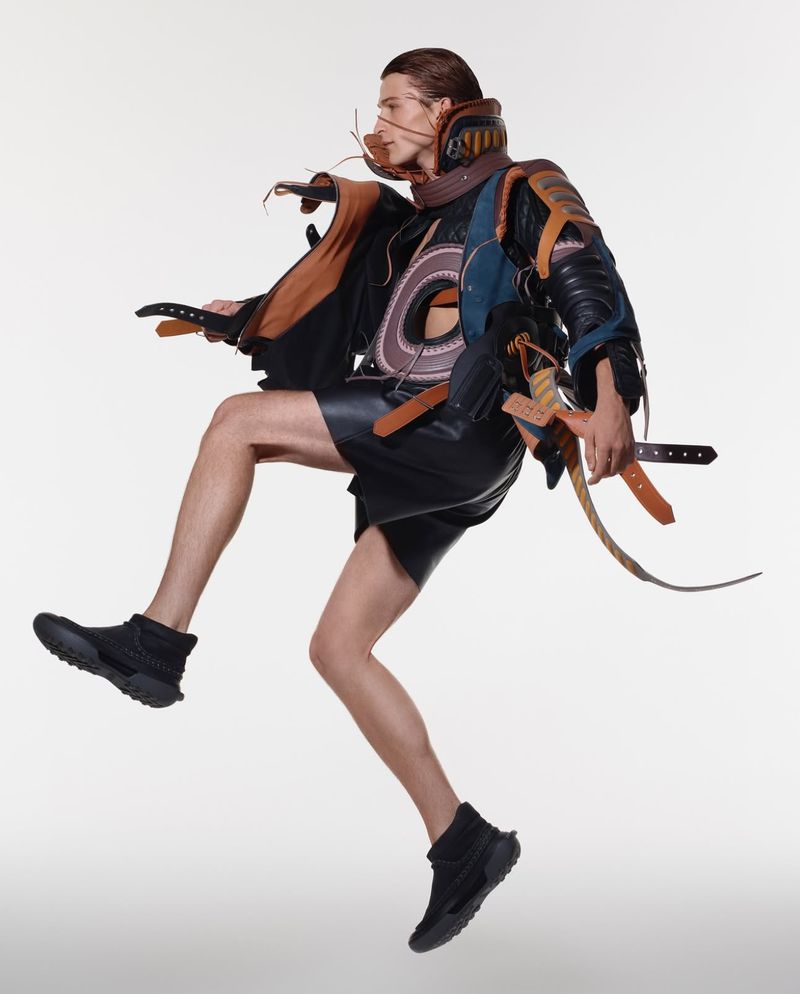 A fair-skinned male fashion model leaps in a photo studio wearing a black jacket, shorts, boots, and an elaborate ornamental harness.