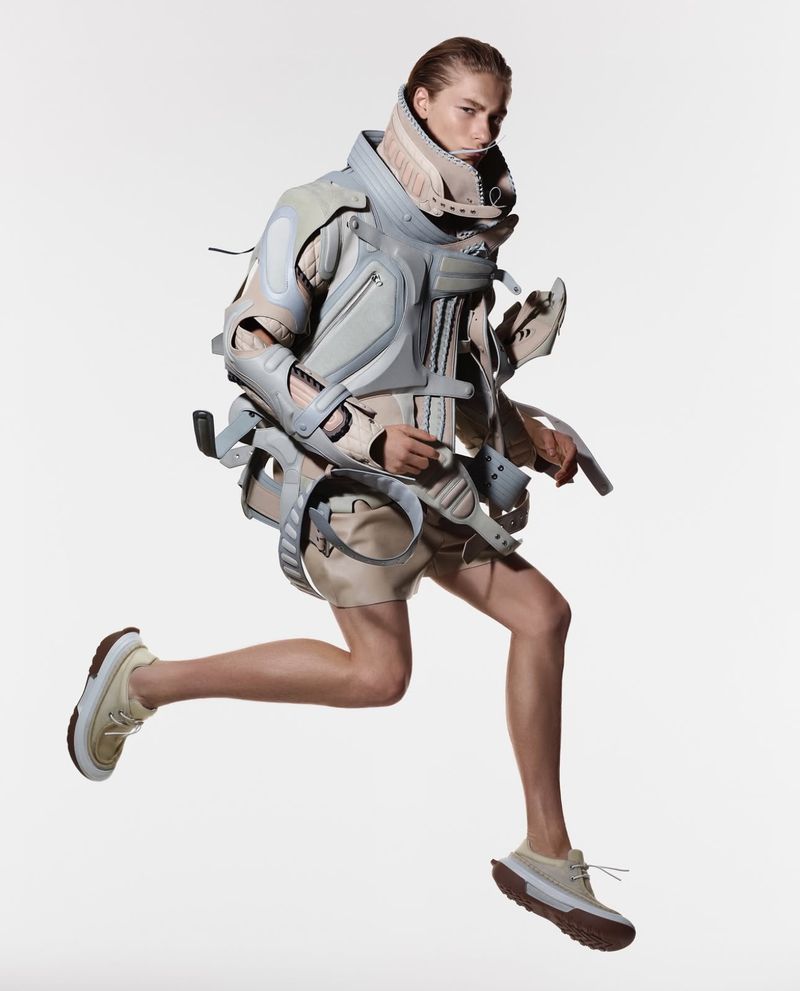 Fashion shot of a person in grey and beige protective gear jumping mid-air against a white background in a studio setting. 