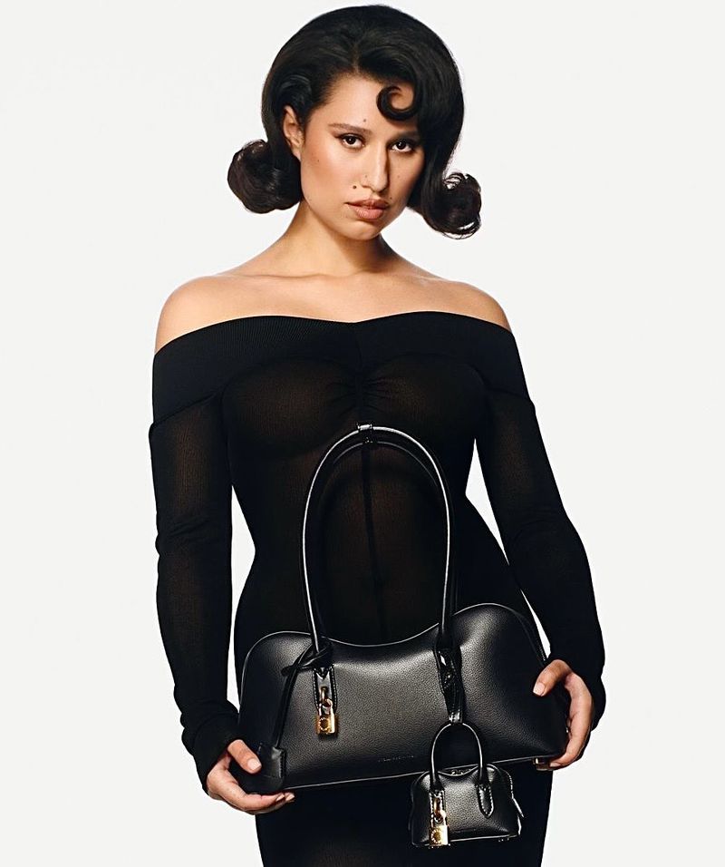 Brunette woman in sheer black dress holding black leather handbag with a mini-bag charm, posing in front of a white background.