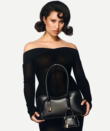 Brunette woman in sheer black dress holding black leather handbag with a mini-bag charm, posing in front of a white background.