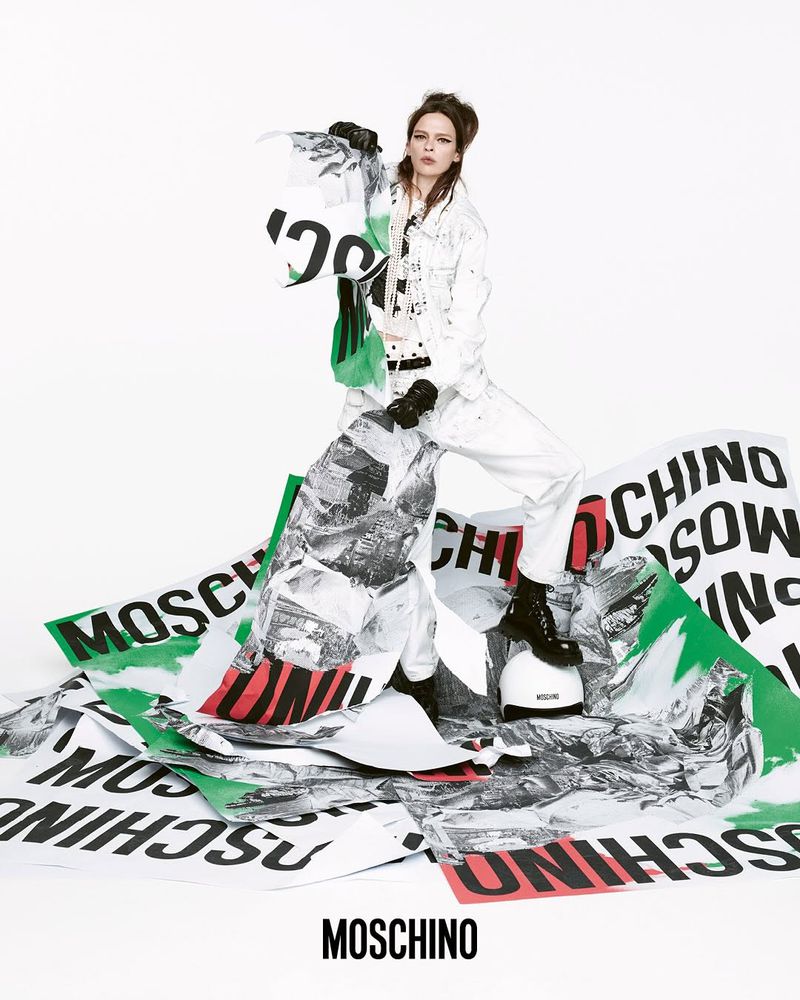 A model poses in a white outfit surrounded by MOSCHINO-branded paper. She is captured indoors with bright lighting, showcasing a high-fashion aesthetic.