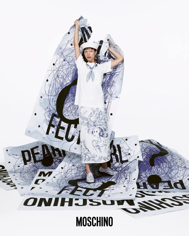Fashion shot of a woman in a sailor hat and clothing, surrounded by oversized posters with graffiti-style text and black scribbles.
