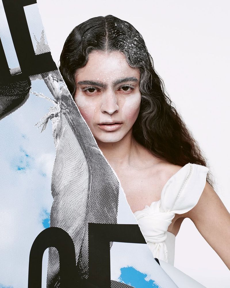 Portrait of a woman with dark wavy hair, styled with white powder and graphic overlay, against a white backdrop in a studio environment.