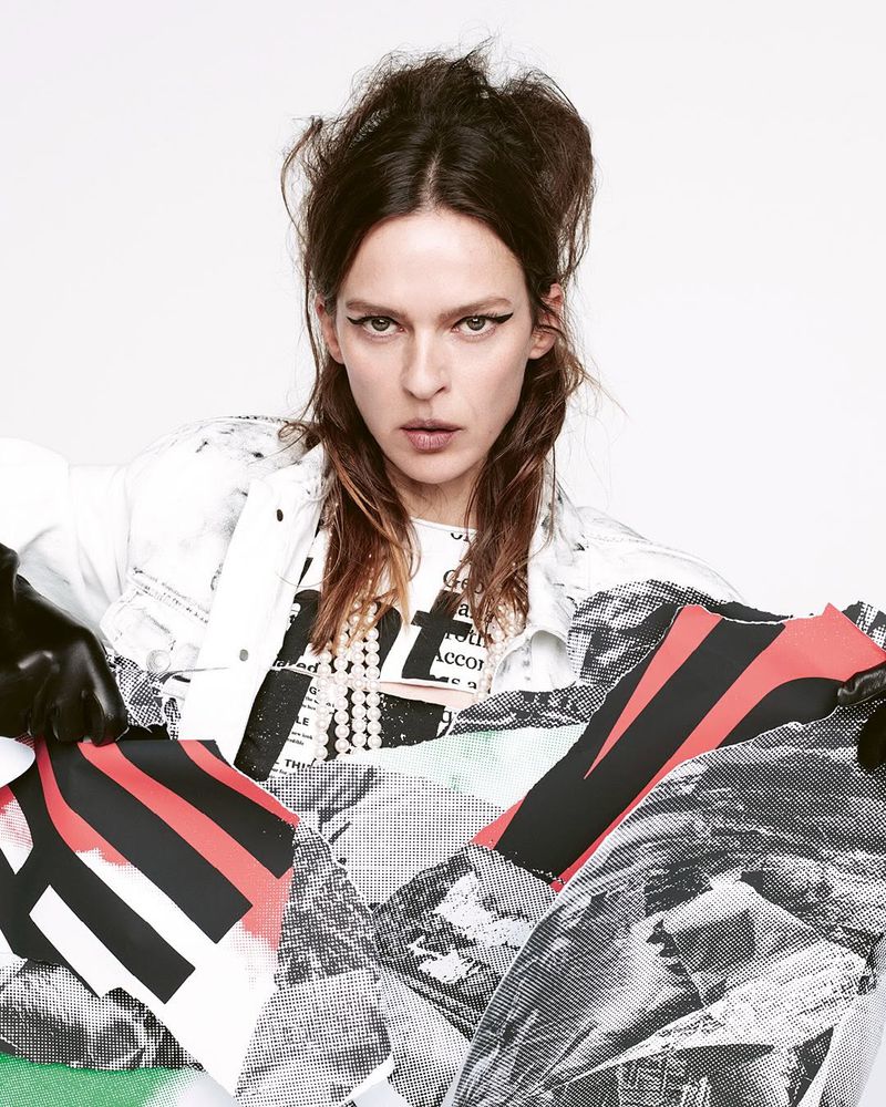 Adult woman poses indoors wearing white jacket, graphic t-shirt, black gloves, and abstract-print tights in a studio shot.
