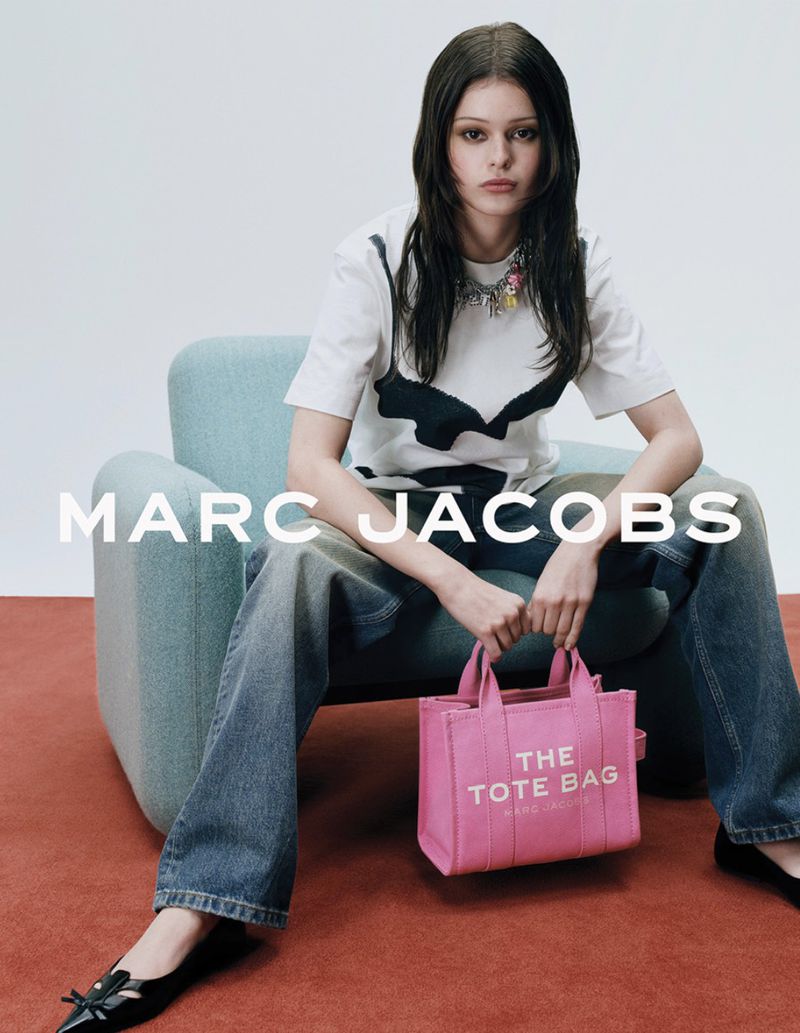 Young woman poses seated holding a pink tote bag in a studio setting with a modern chair, stylishly dressed in casual chic attire.