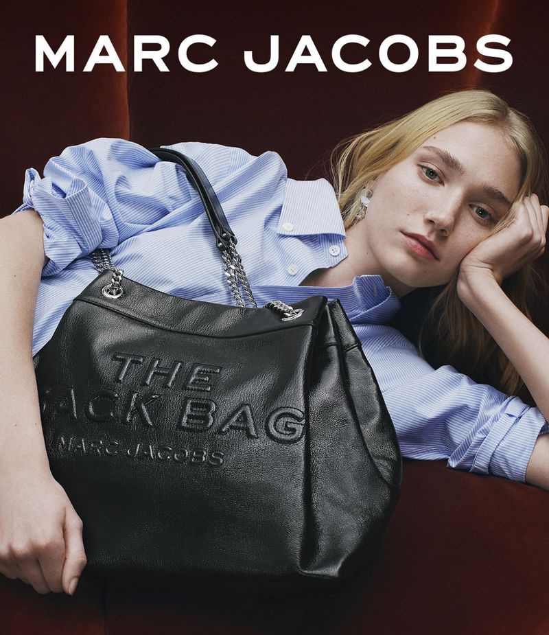 Blonde woman lounges beside a black leather 'The Sack Bag' purse by Marc Jacobs, in a fashion setting against a reddish-brown backdrop.