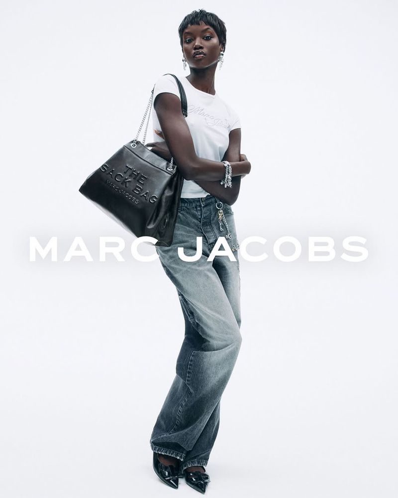 Fashion shot of a Black woman with short hair wearing a white top, blue jeans, and black shoes, carrying a black leather tote bag.