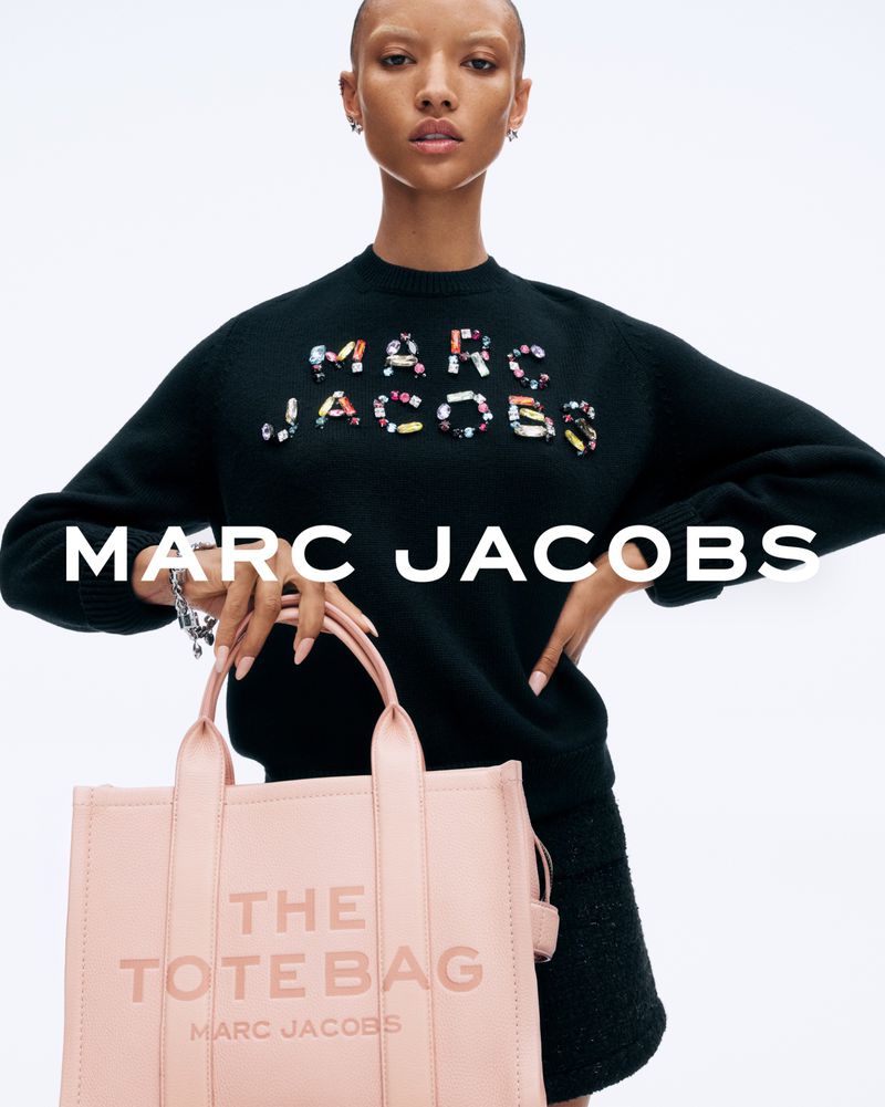 A model poses in a studio wearing a black sweater with 'Marc Jacobs' in colorful beaded letters, holding a pink Marc Jacobs tote bag.