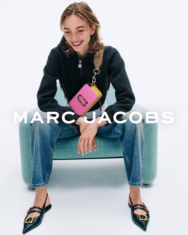 A cheerful woman poses in a black hoodie and jeans while seated, showcasing a pink and mustard-yellow Marc Jacobs handbag.