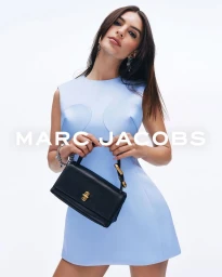 Model Emily Ratajkowski wearing a light blue dress holding a black handbag for Marc Jacobs Spring 2025 campaign against a white backdrop.