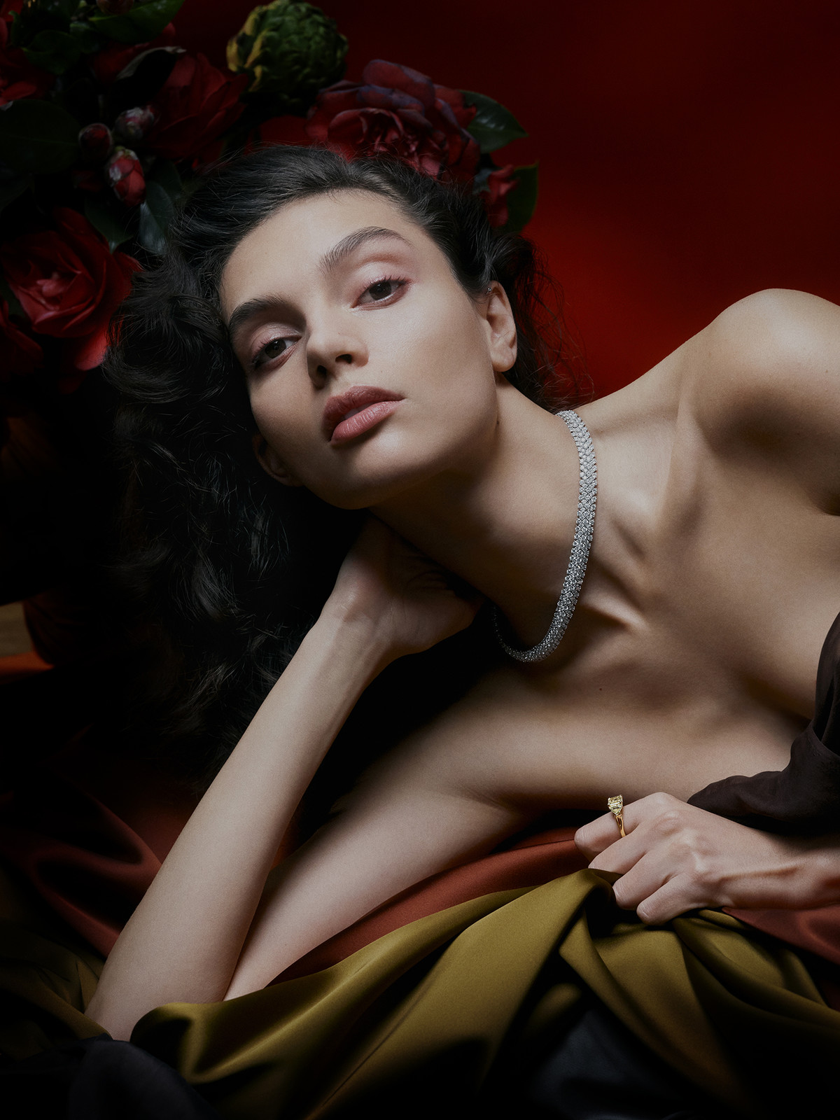 Portrait of a dark-haired woman lying down with a pearl necklace, ring, draped gold fabric, and a floral arrangement on a red background.