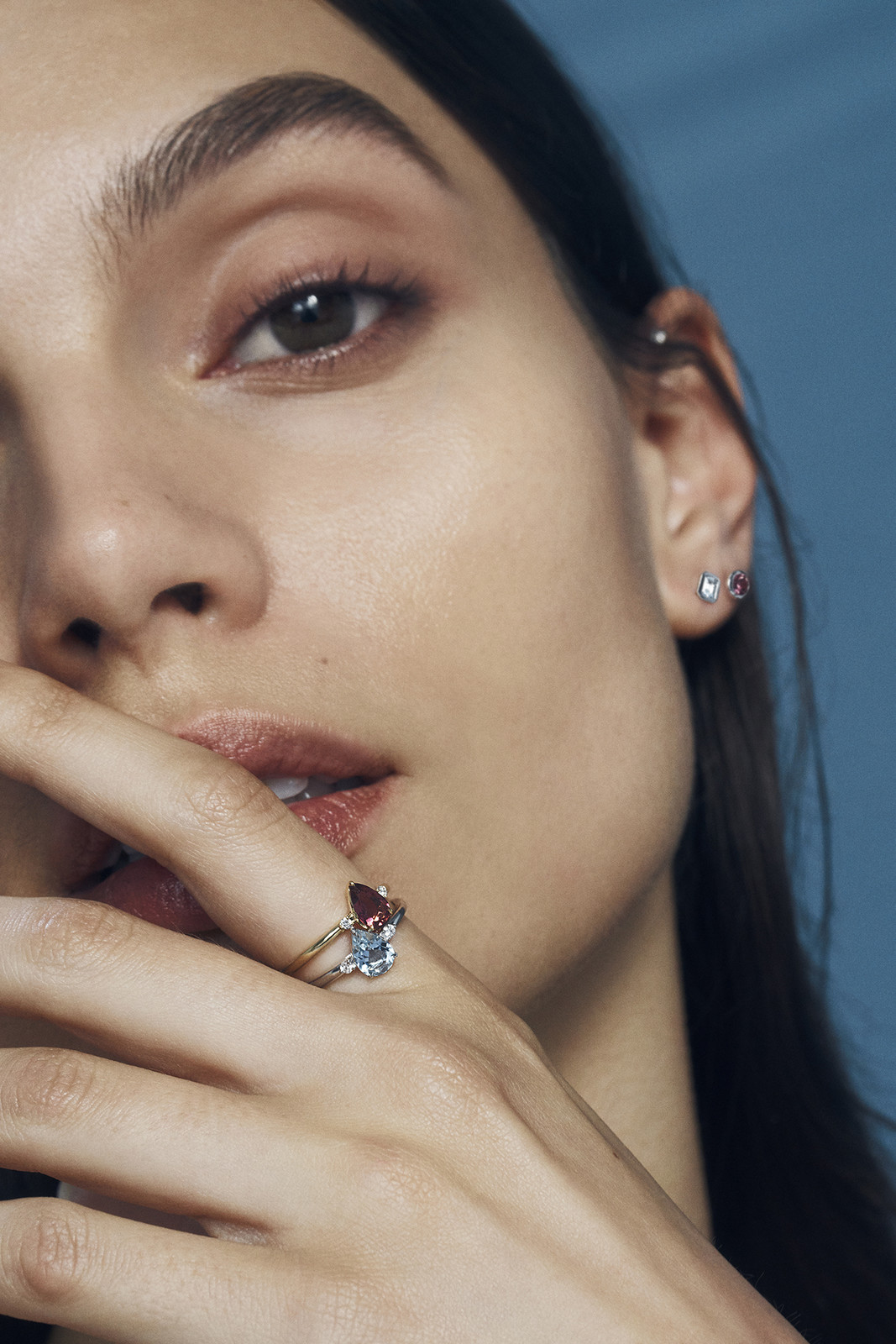 Close-up of a woman wearing a ring with a red stone and a blue stone, posing in front of a blue background in a studio setting.