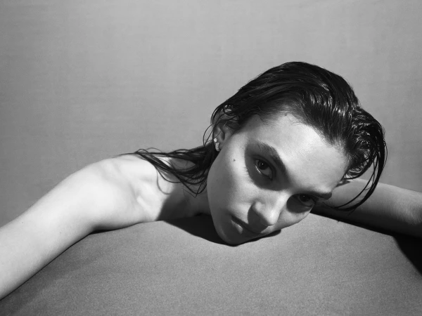 Monochrome portrait of a woman with wet, slicked-back hair, looking directly at the camera while resting on a surface. Studio shot.