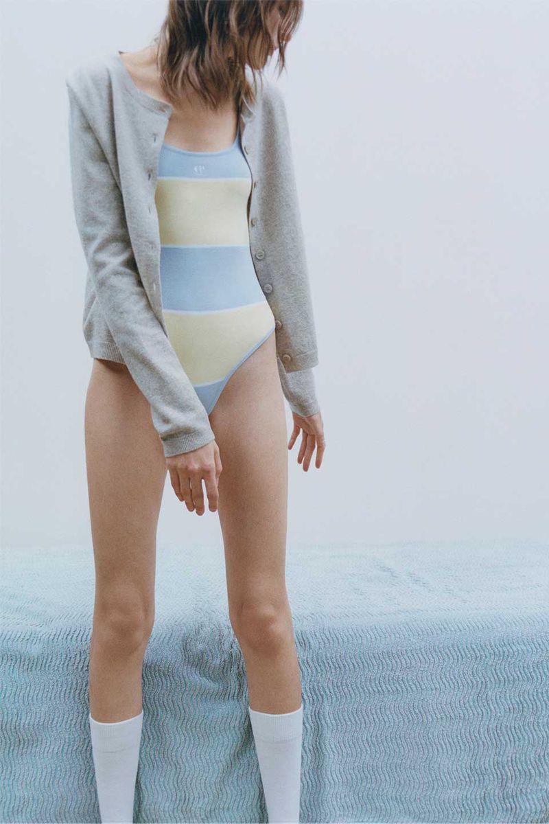 Woman in a striped swimsuit and cardigan stands in a bright studio with a blue textured bedspread. Fashion and lifestyle photography.