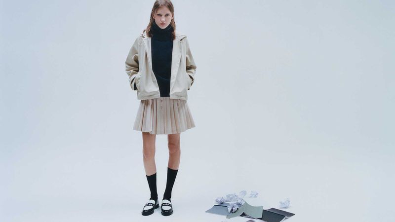 Full-length shot of a young adult female fashion model in a jacket and pleated skirt standing in a bright studio with scattered paper.