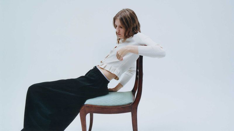 Model wearing white cardigan and black skirt leaning backward on a wooden chair against a plain white background