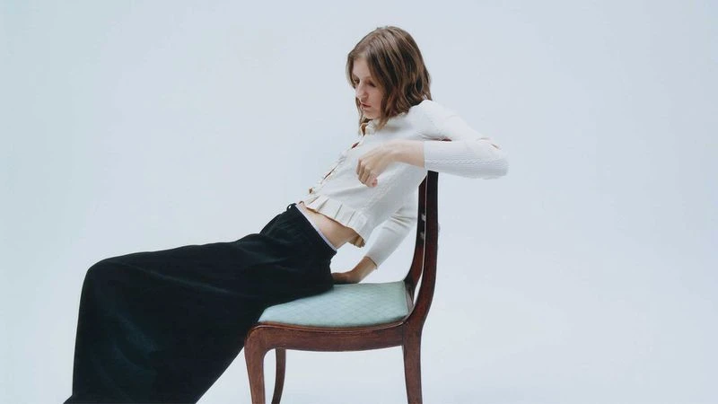 Model wearing white cardigan and black skirt leaning backward on a wooden chair against a plain white background
