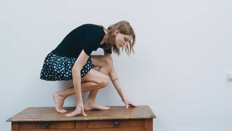Woman crouches on a wooden desk in a minimalist white room, wearing a black top and a patterned black and white skirt.