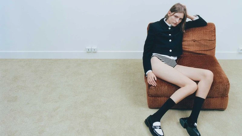 Fashion editorial shot: slender woman in a blazer and underwear strikes a pose while seated on a chair in a minimalist studio.