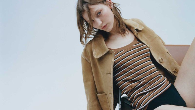 A woman with brown hair gazes downward in a medium shot. She wears a tan jacket and brown striped shirt against a plain background.