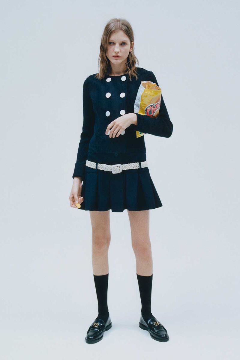 A young woman poses in a preppy navy blue dress, white belt, and black loafers, casually holding a bag of potato chips.