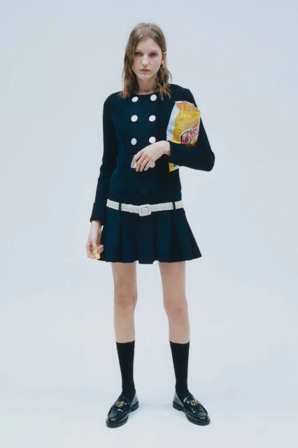 A young woman poses in a preppy navy blue dress, white belt, and black loafers, casually holding a bag of potato chips.