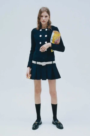 A young woman poses in a preppy navy blue dress, white belt, and black loafers, casually holding a bag of potato chips.
