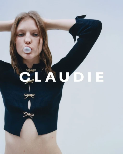 A young woman with long hair is posing indoors against a white background, blowing a bubble with bubblegum while wearing a black top.