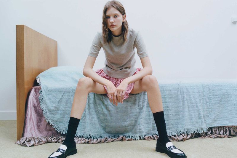 Young woman in a gray shirt and red gingham skirt poses on a bed in black socks and loafers, gazing directly at the camera in a studio setting.