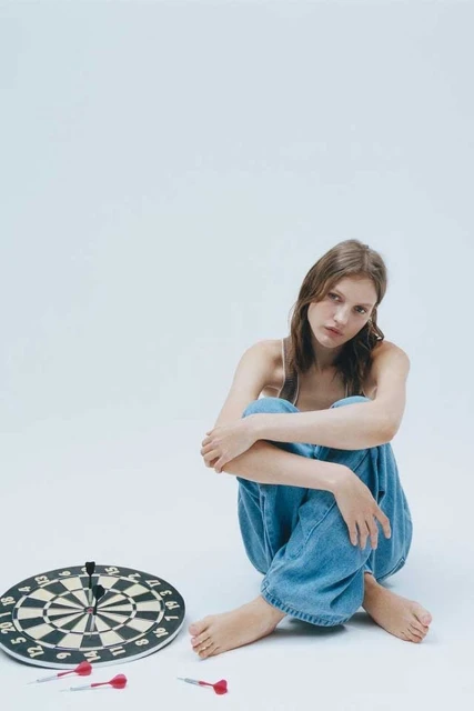 Young woman with long hair sits on a white surface next to a dartboard and darts. Studio shot, portrait orientation.