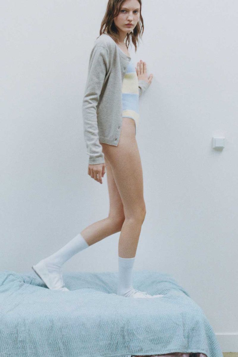 Fashion shot of woman in swimsuit and cardigan standing on a bed with white socks against a white wall in a minimalist style.