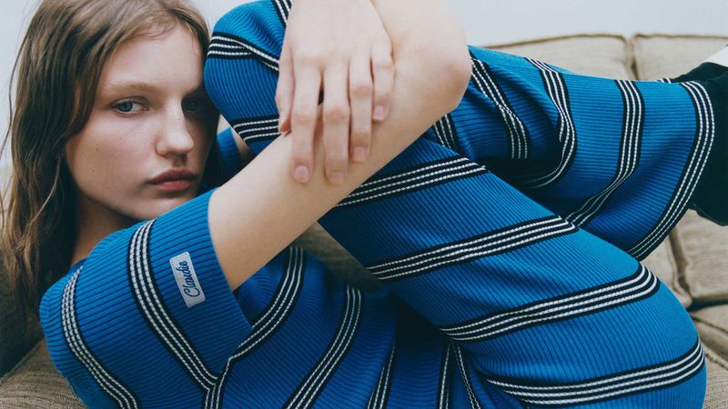Close-up of a young woman with fair skin and blue eyes wearing a blue and white striped top, posing indoors with hands on knees.