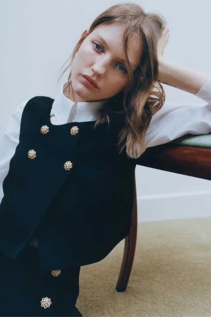A young woman with brown hair poses indoors in a dark vest with gold buttons and a white blouse, leaning on a dark wood chair.