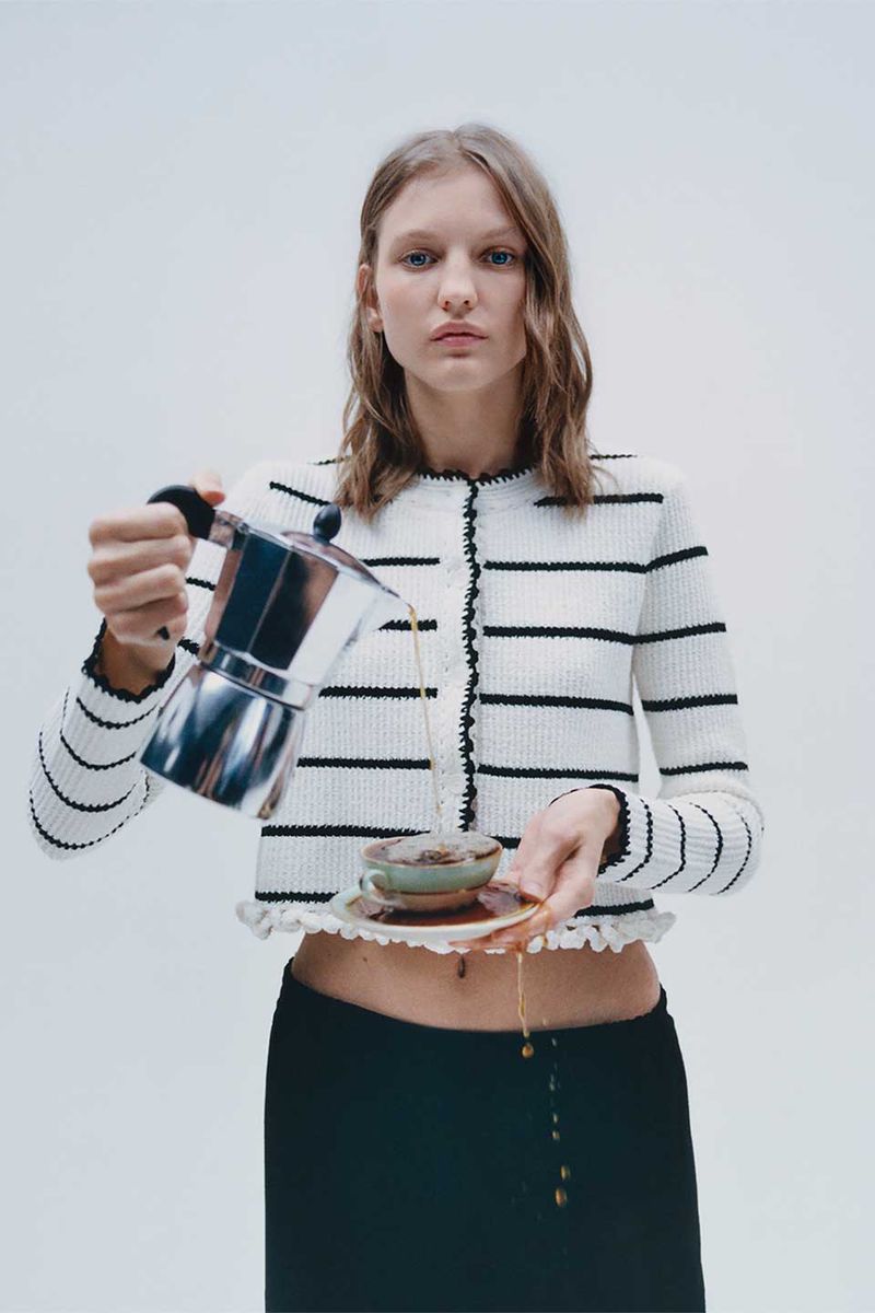 Woman in striped cardigan pours coffee from stovetop pot into a cup on a saucer in a bright photo studio setting.