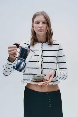 Woman in striped cardigan pours coffee from stovetop pot into a cup on a saucer in a bright photo studio setting.