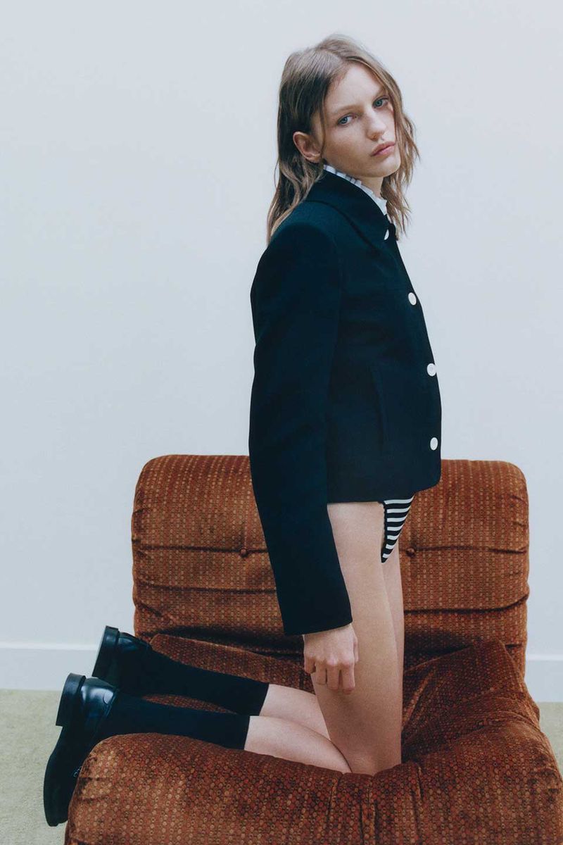 Blonde female fashion model poses on a brown chair, wearing a black jacket, striped bottoms, black socks, and black shoes indoors.