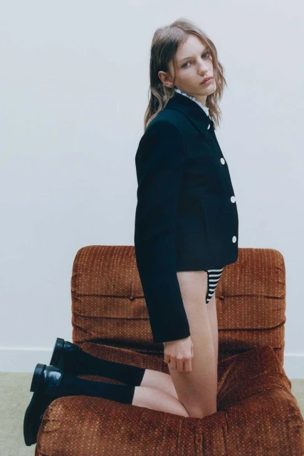 Blonde female fashion model poses on a brown chair, wearing a black jacket, striped bottoms, black socks, and black shoes indoors.
